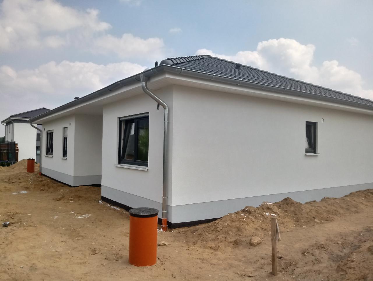 Nowo wybudowany, parterowy dom z białymi ścianami i szarą dachówką, widoczny z zewnątrz, z pomarańczowymi elementami kanalizacji w ziemi.