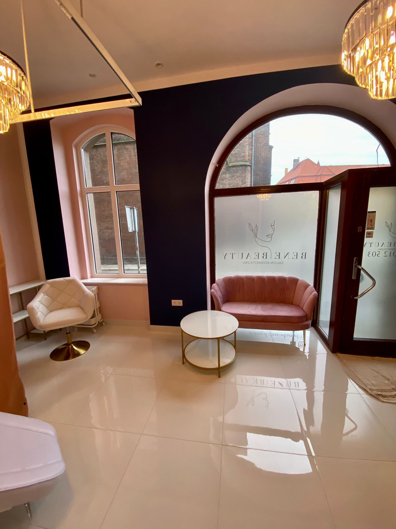 Jasne wnętrze salonu kosmetycznego z różowymi i białymi akcentami, widok na zewnątrz przez zaokrąglone okno i drzwi, lśniąca podłoga z odbiciami.