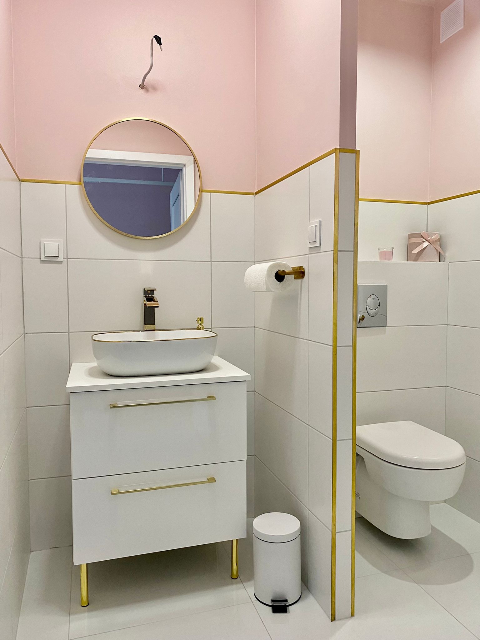 Nowoczesna łazienka w bieli i różu z złotymi akcentami: umywalka nablatowa, okrągłe lustro, biała toaleta i dekoracyjne pudełko na półce.