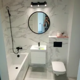 Nowoczesna łazienka z marmurowymi płytkami, czarną armaturą i okrągłym lustrem, w którym odbija się osoba robiąca zdjęcie.