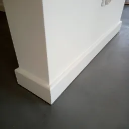 Detal wykończenia wnętrza: biała ściana z listwą przypodłogową, szara podłoga i wystający narożnik z gniazdkiem elektrycznym.