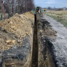 Wąski wykop w ziemi, z widocznym minikoparką i przyczepą w tle, otoczony ziemią i trawą, jesienna sceneria.