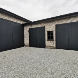 Budynek z trzema czarnymi, metalowymi bramami garażowymi, wykończony jasną cegłą i pokryty szarym żwirem.