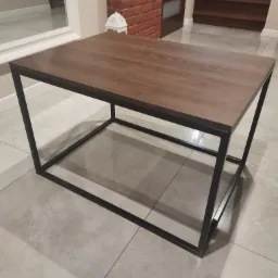 Stolik kawowy z drewnianym blatem i czarną metalową ramą, na tle jasnej podłogi i fragmentu ściany z cegły.