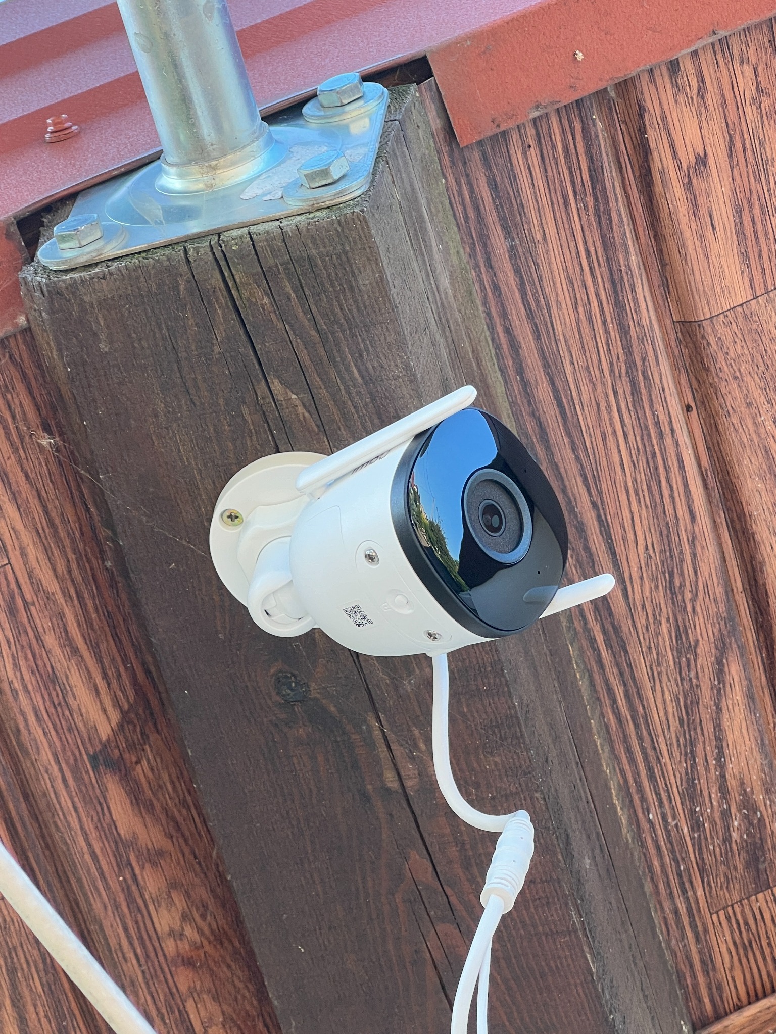 Biała kamera monitoringu zamontowana na drewnianej konstrukcji, z widocznym kablem i elementami mocującymi.