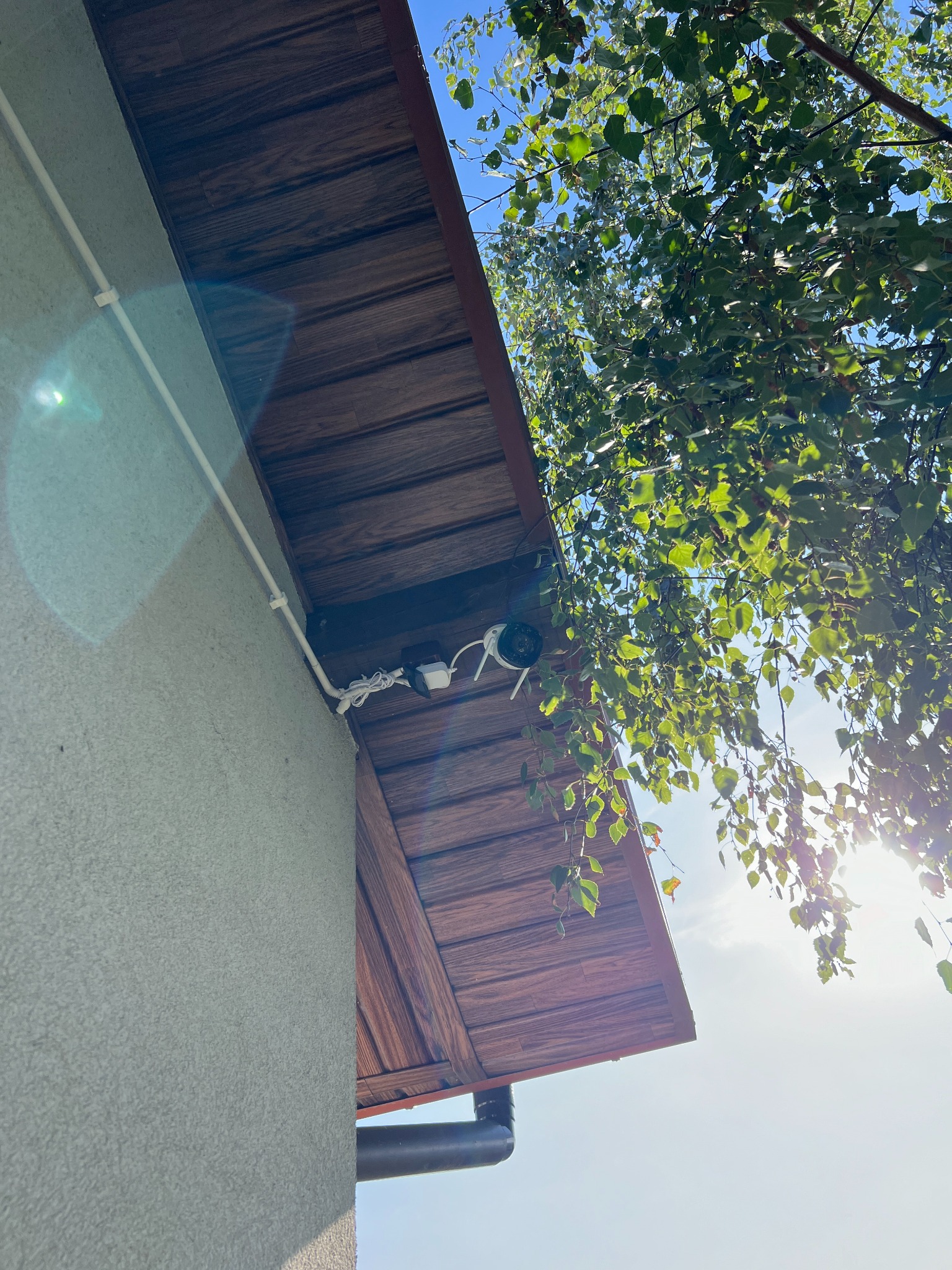 Biała kamera monitoringu zamontowana pod drewnianym okapem dachu, z widocznymi kablami i rurką ochronną, na tle szarej ściany budynku i gałęzi drzewa z liśćmi podświetlonymi słońcem.