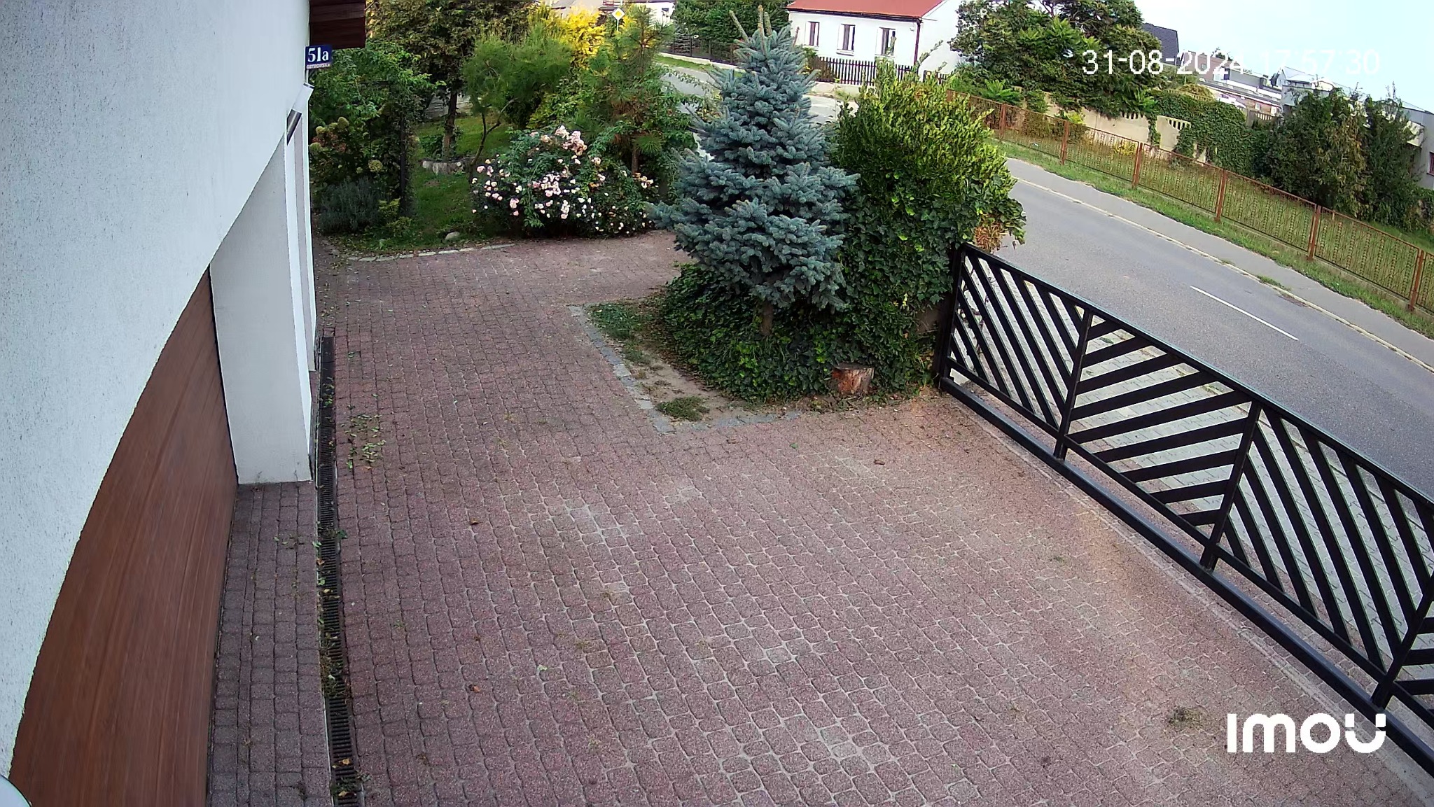 Obraz z kamery monitoringu: podjazd wyłożony kostką, brama z czarnymi, ukośnymi elementami, widok na ulicę i zieleń, data i logo kamery w rogu.