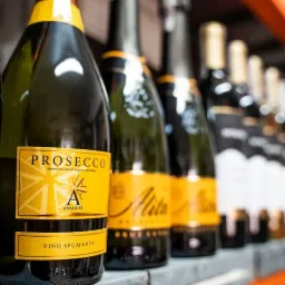 Rząd butelek Prosecco i innych win musujących z żółtymi etykietami na półce w magazynie, widoczne fragmenty regału i inne butelki w tle.