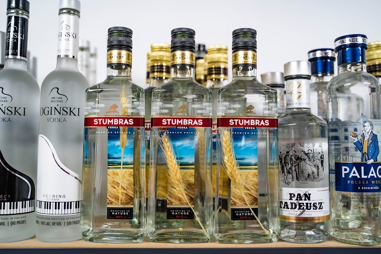 Różne butelki wódki, w tym trzy butelki Stumbras z motywem kłosa zboża na etykiecie, ustawione na półce w hurtowni.