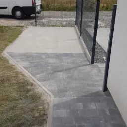 Ułożona ścieżka z kostki brukowej w odcieniach szarości, prowadząca od furtki z czarnej siatki do jasnej elewacji domu, z widocznym fragmentem trawnika i białym samochodem dostawczym w tle.