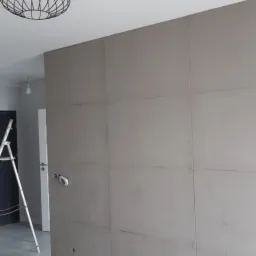 Ściana w trakcie szpachlowania, widoczne łączenia płyt kartonowo-gipsowych, drabina, lampa sufitowa w stylu industrialnym i worki z zaprawą na podłodze.