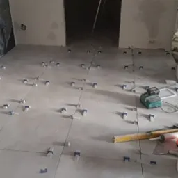 Szare płytki podłogowe w trakcie układania, z plastikowymi krzyżykami dystansowymi, poziomica, szpachelka i wiadro widoczne na podłodze w remontowanym pomieszczeniu.