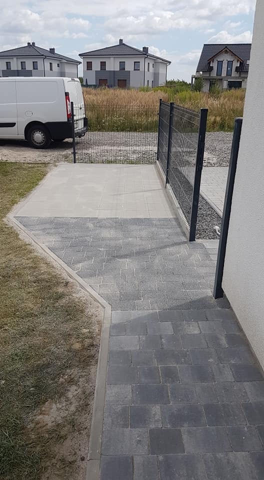 Ułożona ścieżka z kostki brukowej w odcieniach szarości, prowadząca od furtki z czarnej siatki do jasnej elewacji domu, z widocznym fragmentem trawnika i białym samochodem dostawczym w tle.