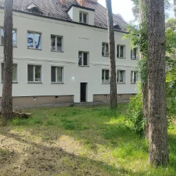 Odnowiona elewacja budynku wielorodzinnego w jasnym kolorze, widoczna przez pnie drzew iglastych na pierwszym planie, z brązowym dachem i ramami okiennymi.