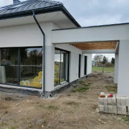 Surowy stan elewacji domu jednorodzinnego z widoczną izolacją w rolkach za oknem, przygotowane bloczki betonowe i robocze rękawice w tle krajobraz wiejski.