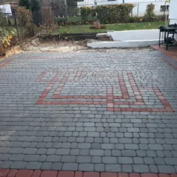 Ułożona nawierzchnia z szarej kostki z czerwoną obwódką i wzorem w kształcie zygzaka, widok na ogród i dom w tle.