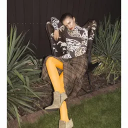 Modelka w bluzce we wzory, spódnicy w kratę i pomarańczowych rajstopach siedzi na krześle z motywem krowy na zewnątrz, z logo 'SALYSE' w rogu.