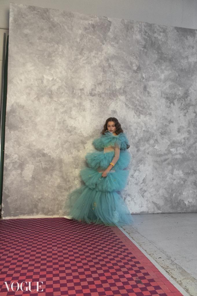 Młoda kobieta w turkusowej, wielowarstwowej sukience z tiulu pozuje na tle szarej ściany w studio Vogue, z czerwono-bordową podłogą w szachownicę.