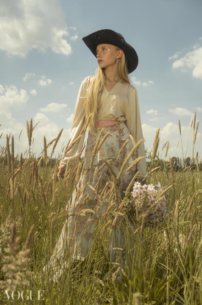 Blondwłosa kobieta w kapeluszu i kwiecistej spódnicy, stojąca na polu trawy z bukietem kwiatów, patrzy w dal pod błękitnym niebem.