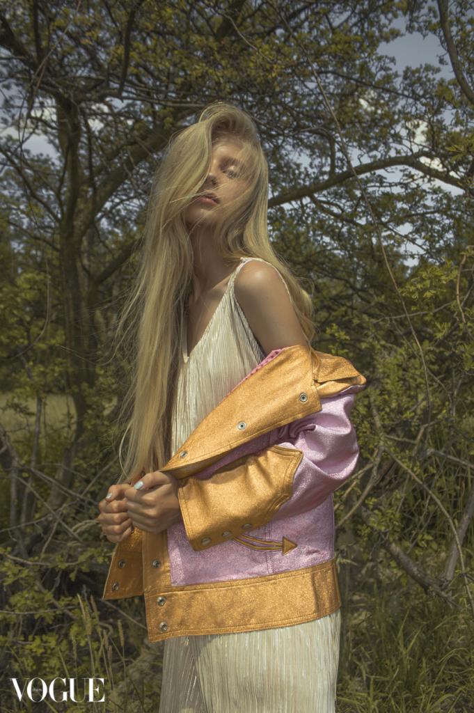 Portret kobiety z długimi blond włosami w plenerze w Zabrzu, ubrana w sukienkę z połyskliwego materiału i kurtkę w odcieniach różu i złota, pozująca na tle drzew.