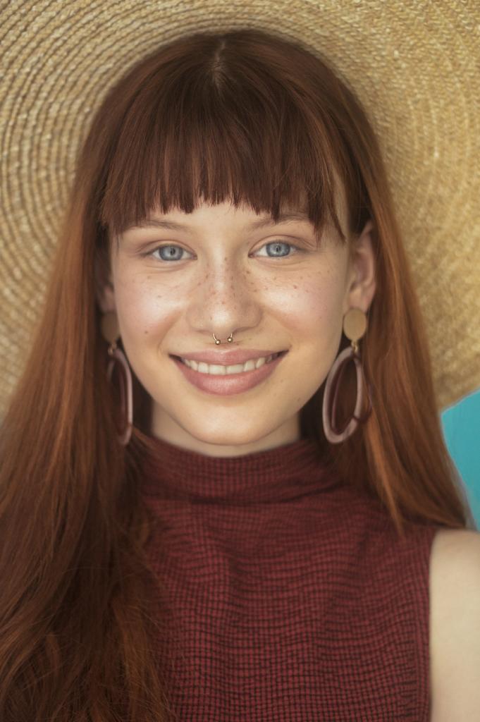 Portret uśmiechniętej rudej kobiety z piegami, w słomkowym kapeluszu, z kolczykami i piercingiem nosa, na tle turkusowej ściany.