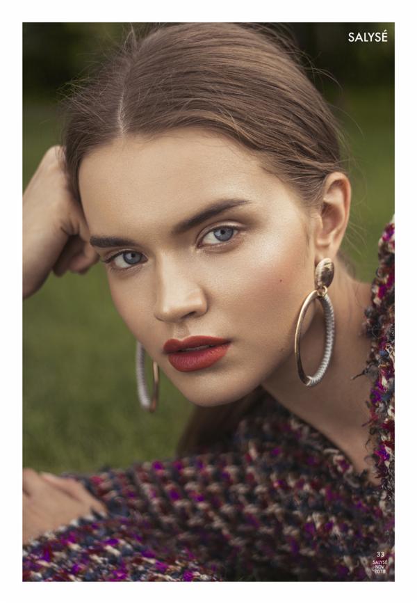 Portret modelki z intensywnym makijażem ust, dużymi, wiszącymi kolczykami i tweedową marynarką na tle zielonej trawy.