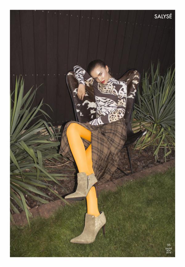 Modelka w bluzce we wzory, spódnicy w kratę i pomarańczowych rajstopach siedzi na krześle z motywem krowy na zewnątrz, z logo 'SALYSE' w rogu.
