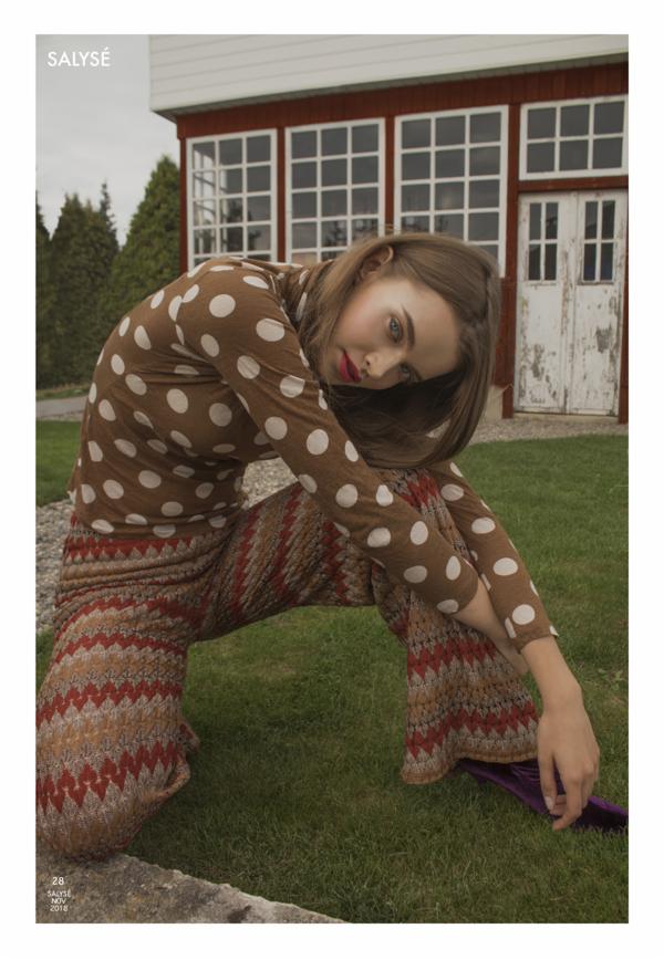 Kobieta w brązowej bluzce w białe kropki i wzorzystych spodniach, siedząca na trawie przed czerwonym budynkiem z białymi oknami i drzwiami