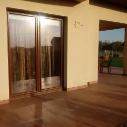 Zamknięte dwuskrzydłowe okno z brązowymi ramami, widok z zewnątrz na taras z brązowymi płytkami i meble ogrodowe w tle.