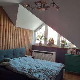 Art Management Sandra Fras - Pokój na poddaszu z drewnianą ścianą z pionowych paneli, łóżkiem z niebieskim zagłówkiem i nakrapianą pościelą, oraz oknem dachowym z trzema doniczkami z roślinami, oświetlonym przez industrialną...