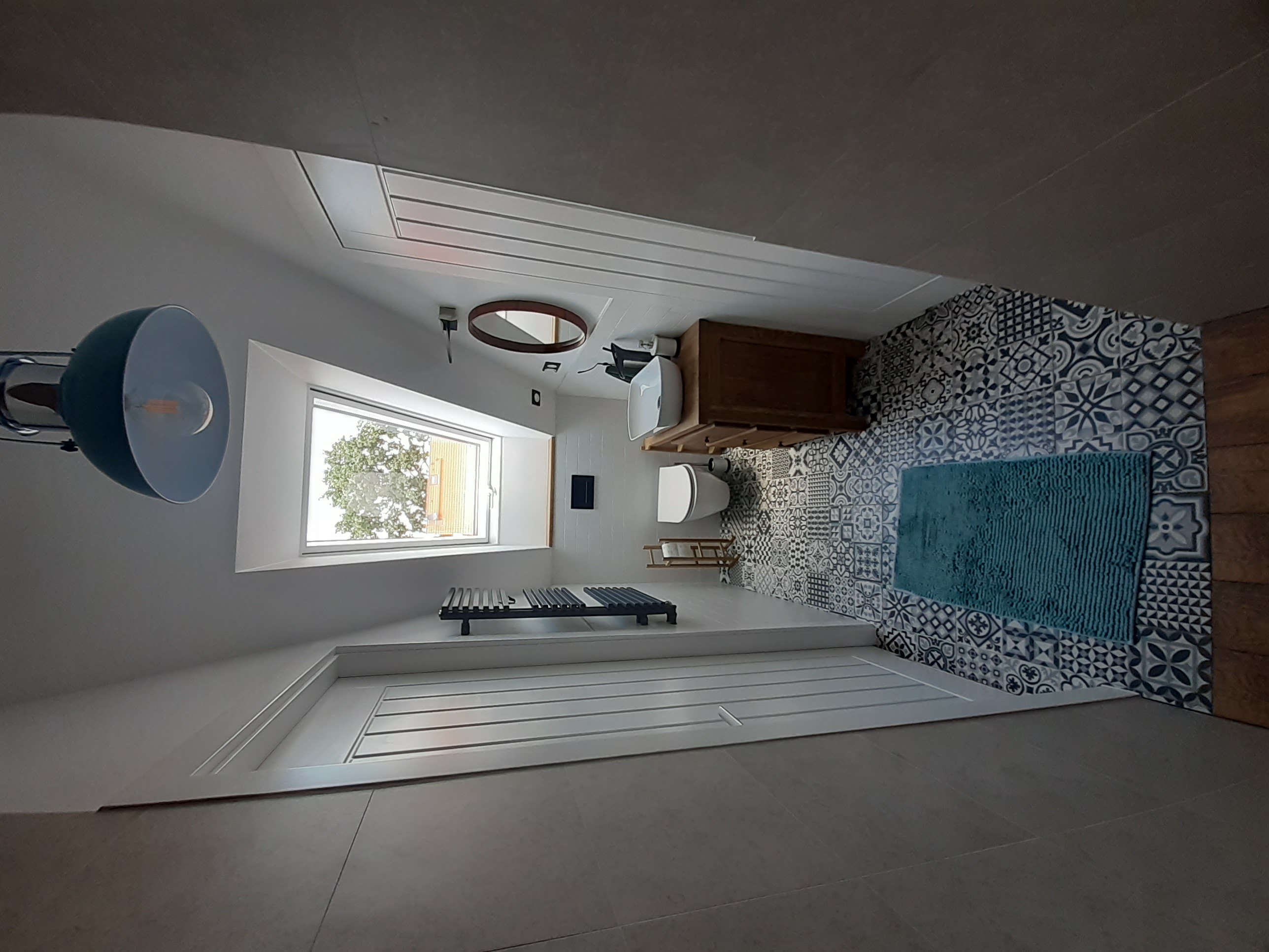 Nowoczesna łazienka na poddaszu z oknem dachowym, wzorzystymi płytkami, drewnianą szafką pod umywalką i niebieskim dywanikiem.