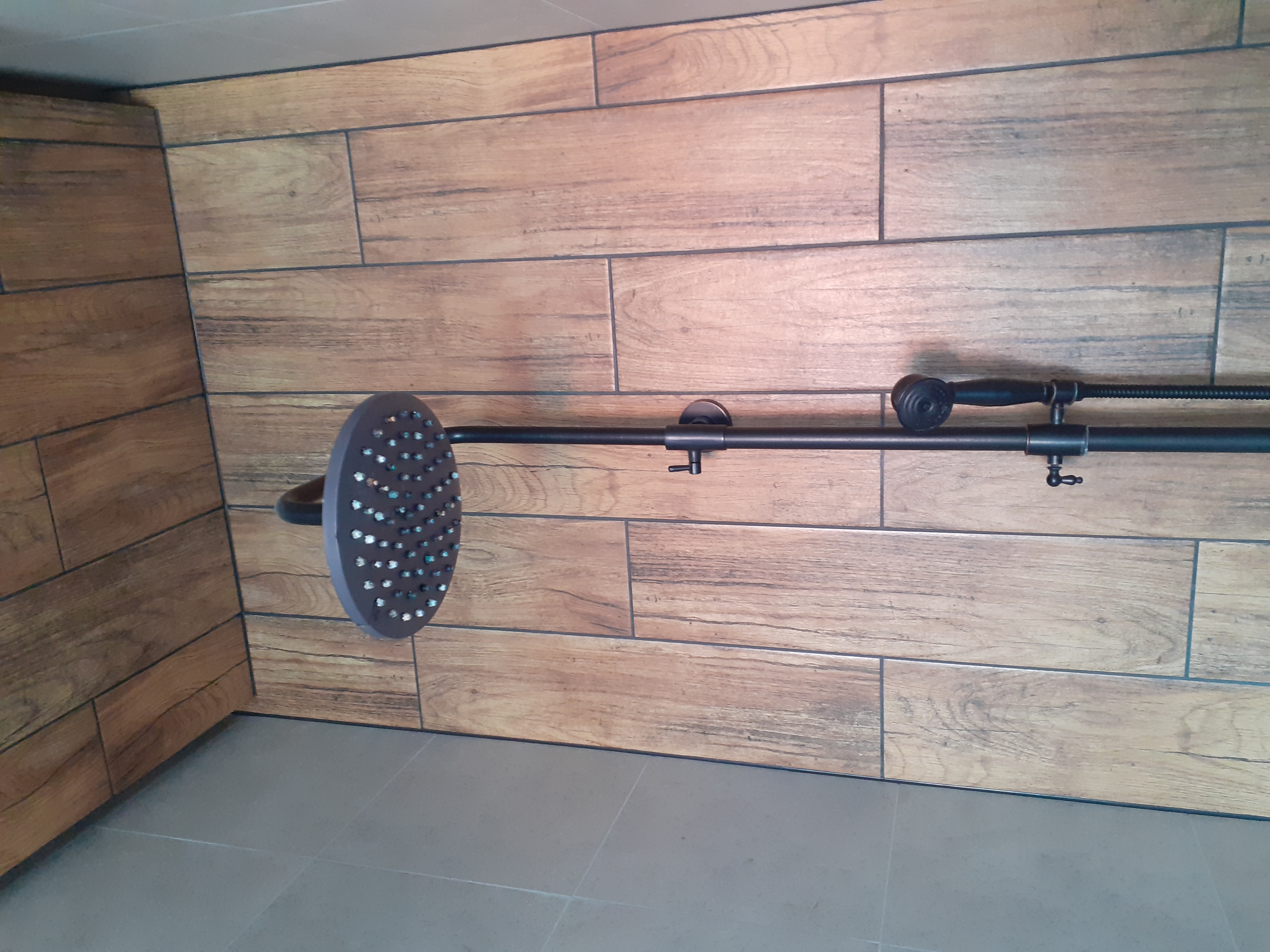 Nowoczesny zestaw prysznicowy w kolorze ciemnego brązu z dużą deszczownicą i dodatkową słuchawką, zamontowany na ścianie wyłożonej płytkami imitującymi drewno w łazience.