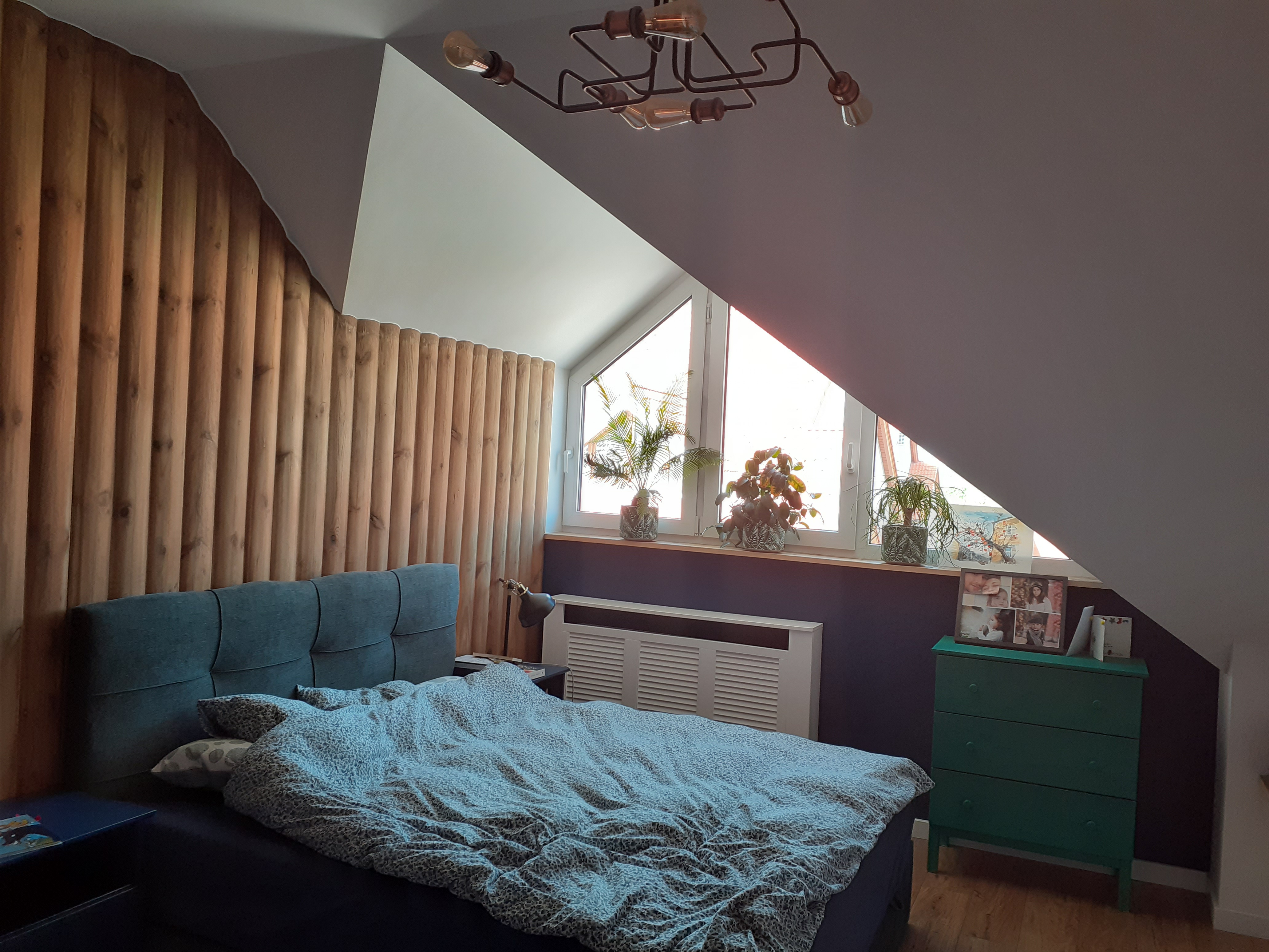 Pokój na poddaszu z drewnianą ścianą z pionowych paneli, łóżkiem z niebieskim zagłówkiem i nakrapianą pościelą, oraz oknem dachowym z trzema doniczkami z roślinami, oświetlonym przez industrialną...