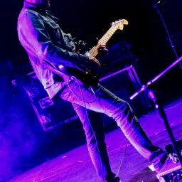 Gitarzysta w jeansowej kurtce gra na gitarze elektrycznej na scenie oświetlonej fioletowym światłem, z widocznymi efektami pod nogami i mikrofonem.