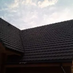 Ciemny, metalowy dach z widoczną teksturą dachówki na drewnianym domu, sfotografowany pod lekkim kątem w pochmurny dzień.