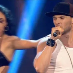 Wokalista w czapce i białym podkoszulku śpiewa do mikrofonu na scenie, obok tancerka w czarnej cekinowej sukience, scena oświetlona niebieskim światłem.