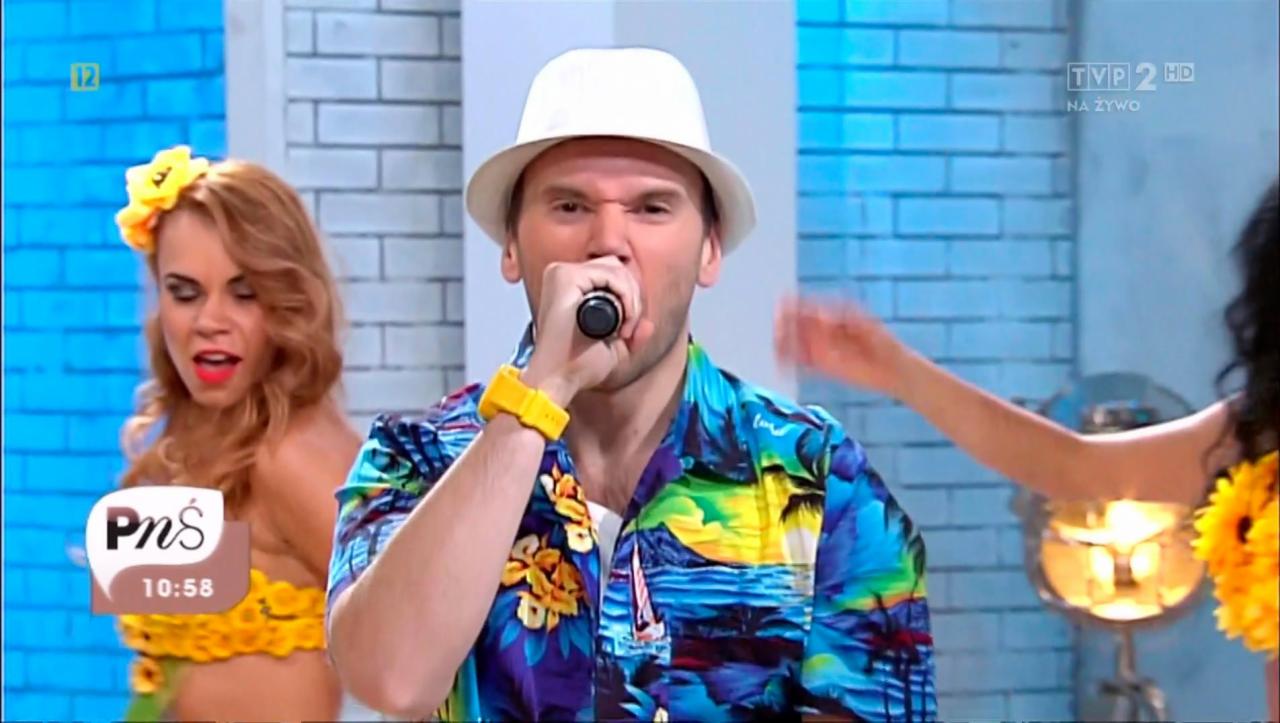 Wokalista w hawajskiej koszuli i białym kapeluszu śpiewa do mikrofonu, w tle dwie tancerki w strojach z motywem słoneczników, występ w programie telewizyjnym na tle niebieskiej ceglanej ściany.