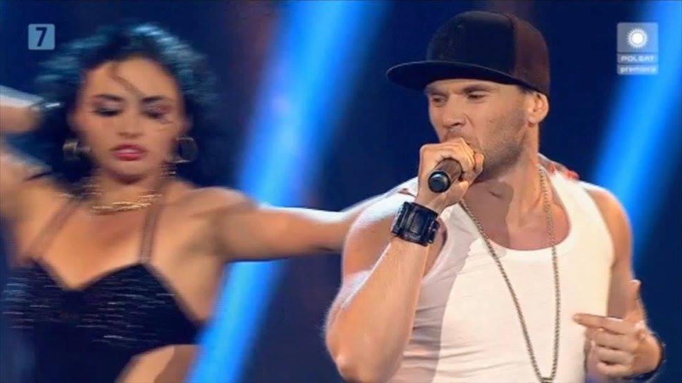 Wokalista w czapce i białym podkoszulku śpiewa do mikrofonu na scenie, obok tancerka w czarnej cekinowej sukience, scena oświetlona niebieskim światłem.