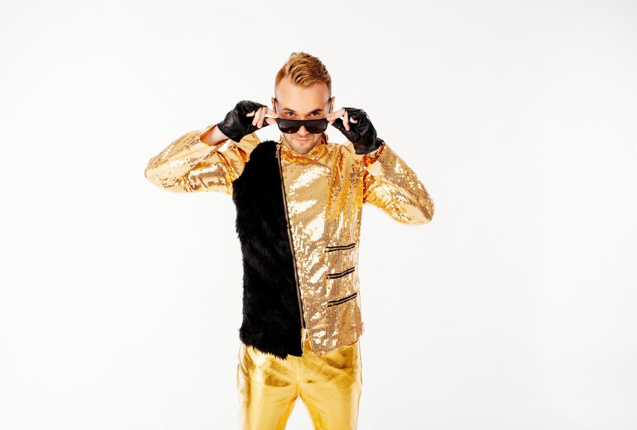 Mężczyzna w błyszczącej złotej kurtce z czarnym futrzanym panelem i złotych spodniach, poprawia okulary przeciwsłoneczne na białym tle.