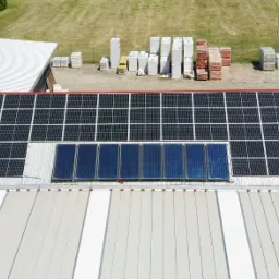 Widok z góry na instalację paneli słonecznych i kolektorów słonecznych na dachu budynku przemysłowego, z logo firmy Voltero widocznym na dachu, oraz składowanymi materiałami budowlanymi w tle.