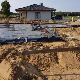 Fundamenty domu jednorodzinnego w trakcie budowy: zbrojenie, folia izolacyjna, rury kanalizacyjne, otoczenie placu budowy z piaskiem i częściowo ukończonym domem w tle.