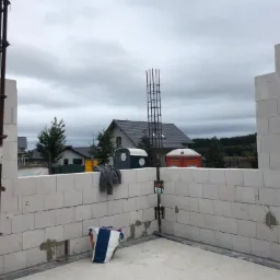 Wznoszenie ścian z bloczków betonu komórkowego na budowie domu jednorodzinnego; widoczne zbrojenie pionowe, tymczasowe toalety w tle.