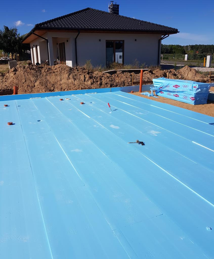 Błękitna izolacja termiczna fundamentów z widocznym nowym domem w tle, przygotowana do dalszych prac budowlanych.