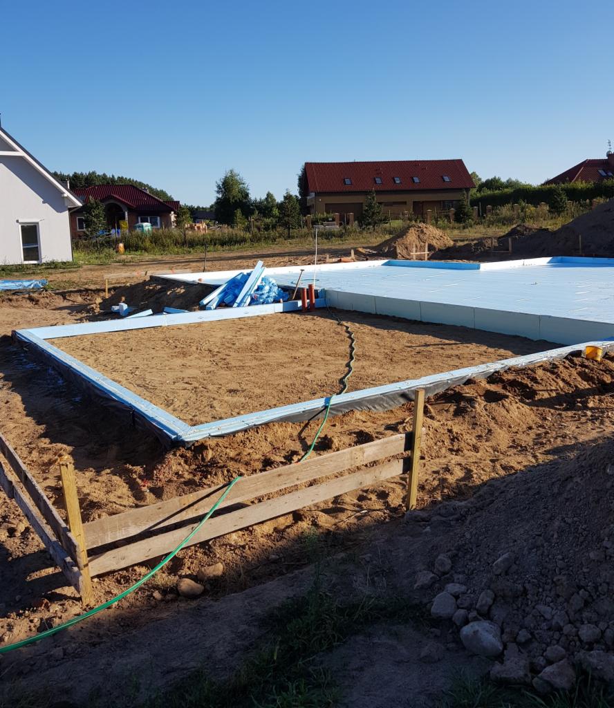 Przygotowanie terenu pod budowę domu w Szczecinie: wykopane fundamenty, szalunki drewniane, izolacja termiczna z niebieskiego styropianu, widok na okoliczne domy.