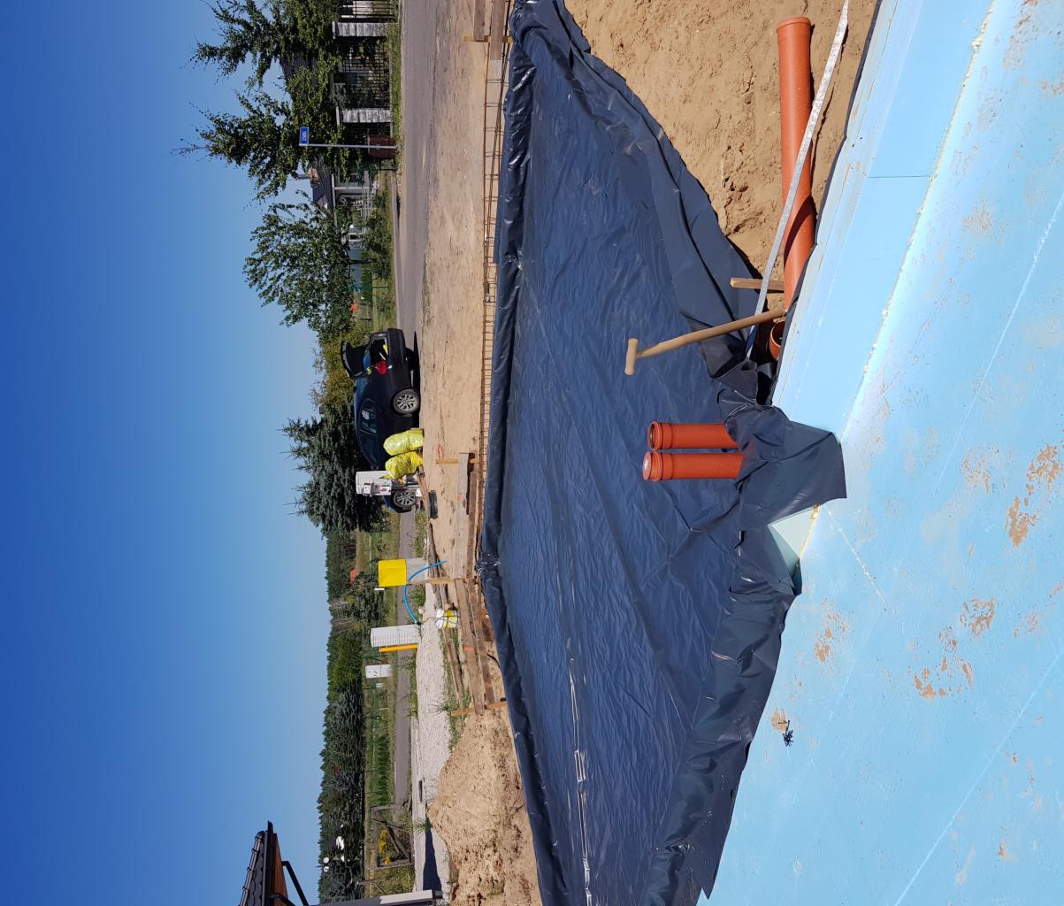 Przygotowanie podłoża pod fundamenty: izolacja folią, rury kanalizacyjne, szalunek, ocieplenie styropianem, widoczne narzędzia i samochód na tle krajobrazu.