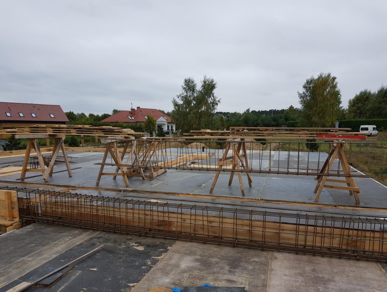 Zbrojenie fundamentów domu jednorodzinnego na przygotowanej płycie, widoczne drewniane podpory i szalunki, w tle zabudowa mieszkalna i zalesiony krajobraz.