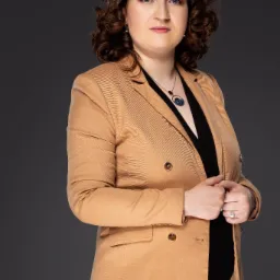 Portret kobiety w beżowej marynarce, czarnej bluzce i ozdobnym naszyjniku, z delikatnym uśmiechem na twarzy, na jednolitym szarym tle.