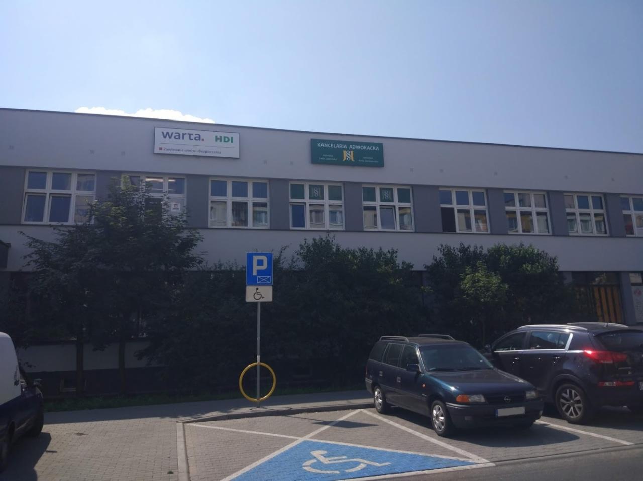Budynek z szyldami Warta HDI i Kancelaria Adwokacka nad parkingiem z miejscem dla niepełnosprawnych i zaparkowanymi samochodami.