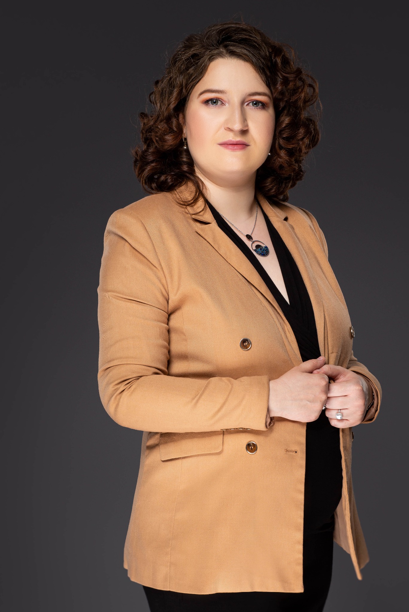 Portret kobiety w beżowej marynarce, czarnej bluzce i ozdobnym naszyjniku, z delikatnym uśmiechem na twarzy, na jednolitym szarym tle.