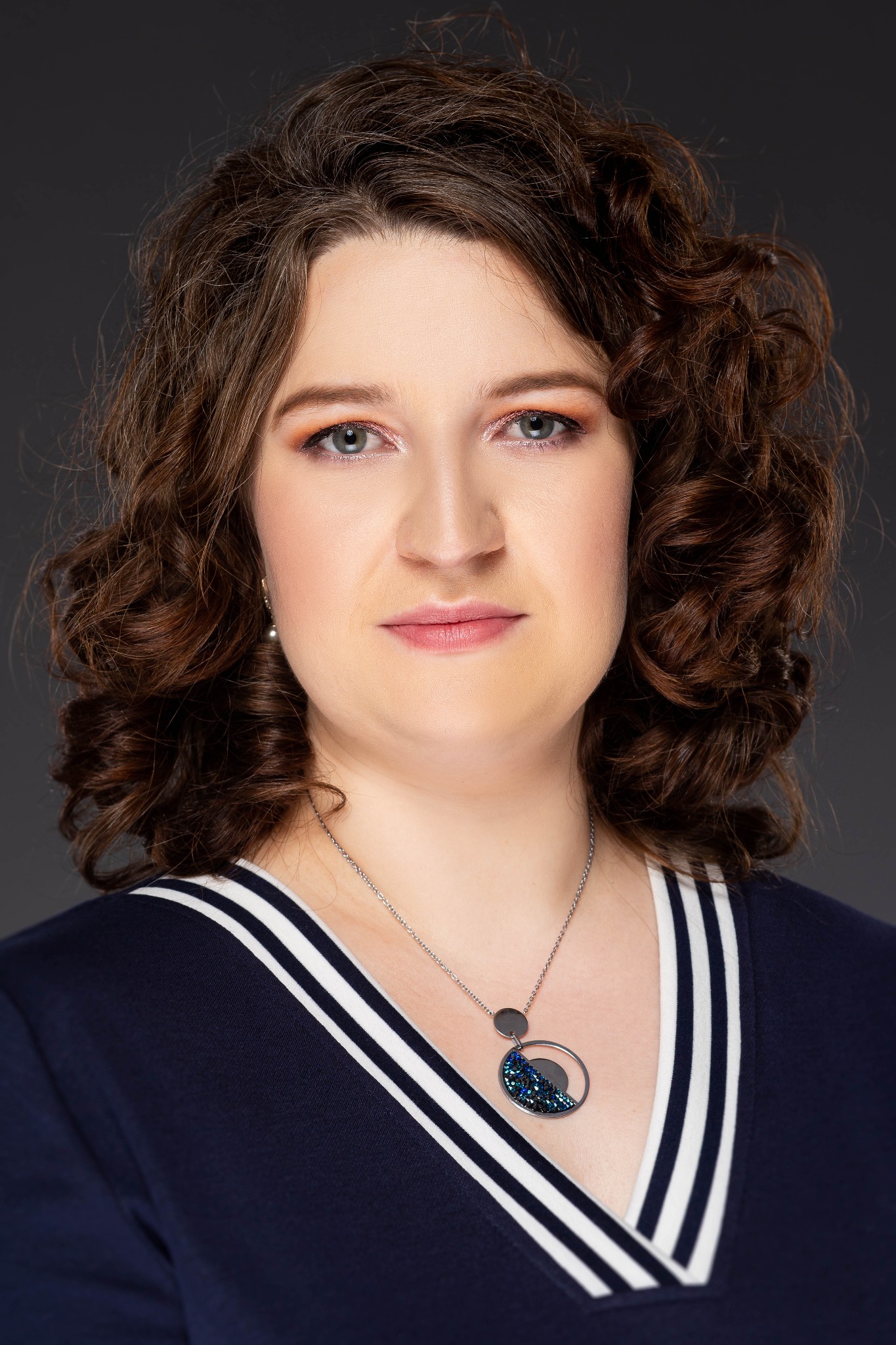 Portret kobiety z kręconymi brązowymi włosami, w granatowej bluzce w białe paski i srebrnym naszyjniku z okrągłą zawieszką z niebieskim brokatem, na ciemnoszarym tle.